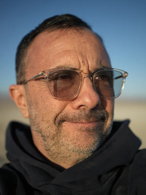 Middle-aged man, close-cropped beard, clear-framed tinted glasses, dark hoodie, photographed outdoors in an arid open la